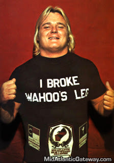 A man with long blonde hair wearing a black retro t-shirt that says "I Broke Wahoo's Leg" with the Mid-Atlantic championship belt referencing the wrestling competition in the 1970s in which Greg Valentine won.
