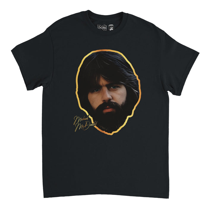 A retro, souvenir black short sleeved t-shirt that says "Michael McDonald" in gold text with an image of the iconic singer and songwriter's face, framed by his dark black brown thick hair and beard and surrounded by a gold outline. Known for being the lead vocalist of the Doobie Brothers rock band and a backing vocalist for Steely Dan.