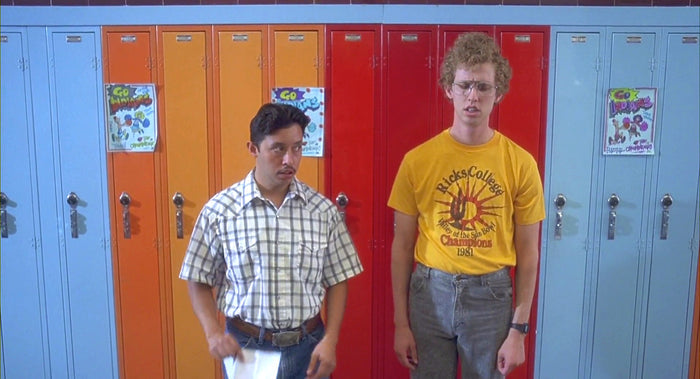 Two men are standing in front of a row of blue, red, and orange lockers, the one on the left wearing a plaid shirt, while the one on the right is wearing a yellow t-shirt with a graphic design that says "Ricks College Valley of the Sun Bowl Champions 1981" with the image of a red, spikey sun. From the movie 'Napoleon's Dynamite' starring Jon Heder as Napoleon Dynamite and Efren Ramirez as Pedro Sánchez.