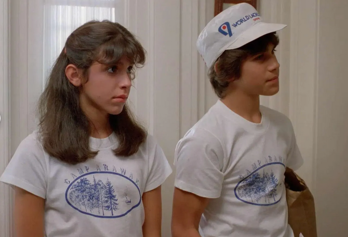 Two people, including Angela Baker played by Felissa Rose and Ricky Thomas played by Jonathan Tiersten, both wearing Camp Arawak t-shirts with images of a forest and lake with a sailboat, the boy wearing a "World Lacross 82" hat from the iconic 1983 horror slasher film "Sleepaway Camp".