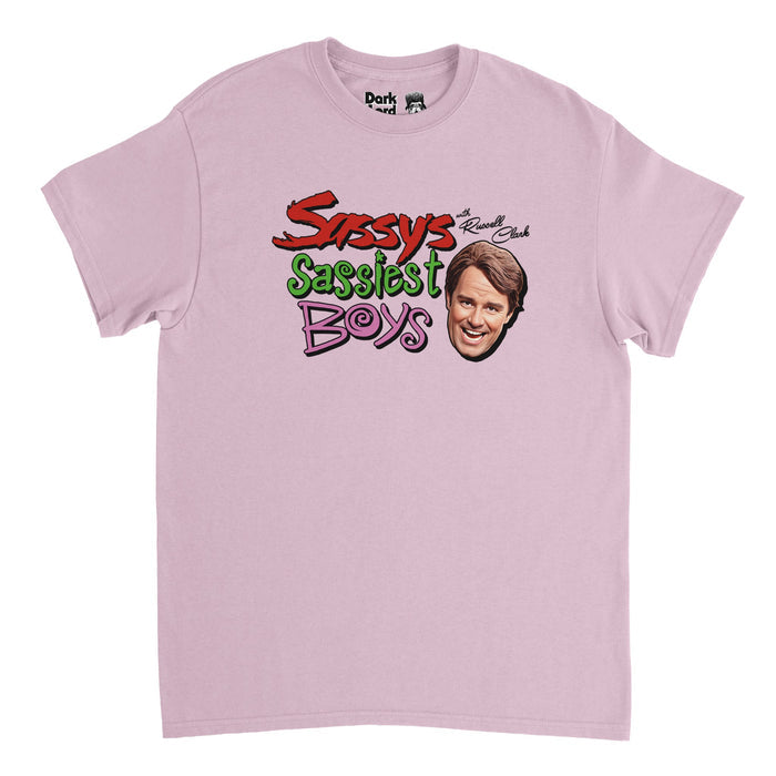 A retro, light pink 70s style short sleeved t-shirt that says "Sassy's Sassiest Boys with Russell Clark" in bold red, bright green, and pink swirly text next to an image of character Russell Clark, played by iconic comedian and actor Phil Hartman, referencing the SNL skit in which he's the senior editor of Sassy Magazine and interviews multiple people including Christian Slater.