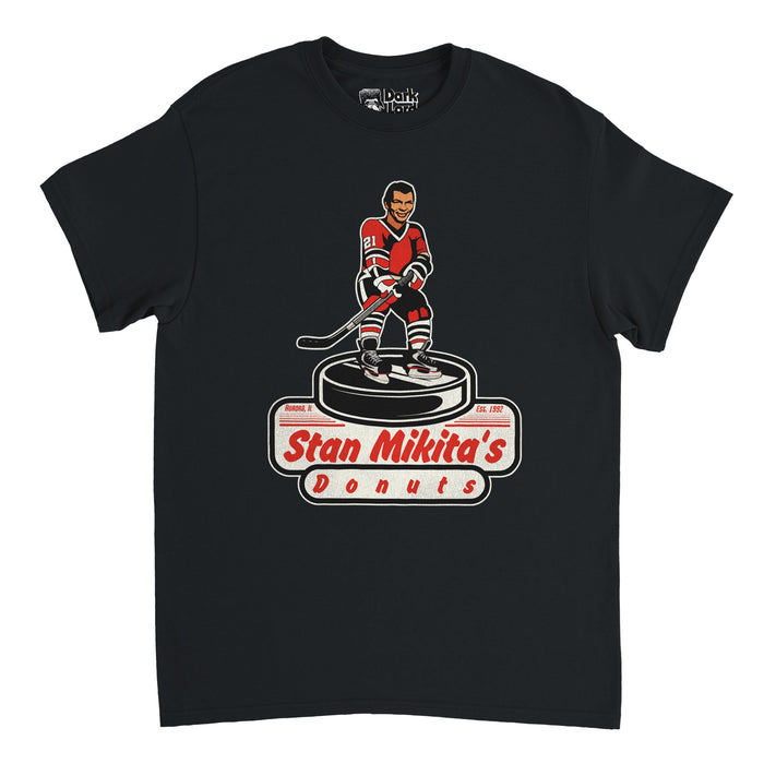 A vintage, souvenir Stan Mikita's Donuts t-shirt in the color black. Includes a graphic of a hockey player wearing a red, black, and white jersey with the number 21, holding a hockey stick and standing on a large hockey puck. Shirt also says Aurora, IL and Est. 1992. References the famous fictional donut shop named after the iconic Chicago Blackhawks hockey player in the pop comedy culture TV show Wayne's World.