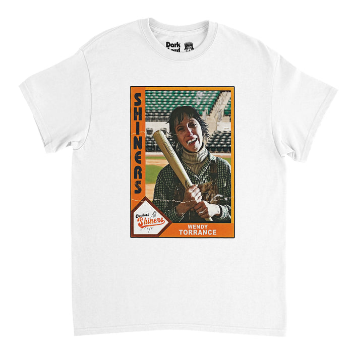 A retro, 70s style white colored t-shirt with a rectangular image, the background orange, that says "Shiners" next to an image of fictional character Wendy Torrance holding a bat and smiling on a baseball field, with a graphic of a home plate that says "Overlook Shiners" with the image of a baseball and the name "Wendy Torrance". References the horror novel and movie "The Shining" by Stephen King and the iconic actress Shelly Duvall who played Wendy Torrance.