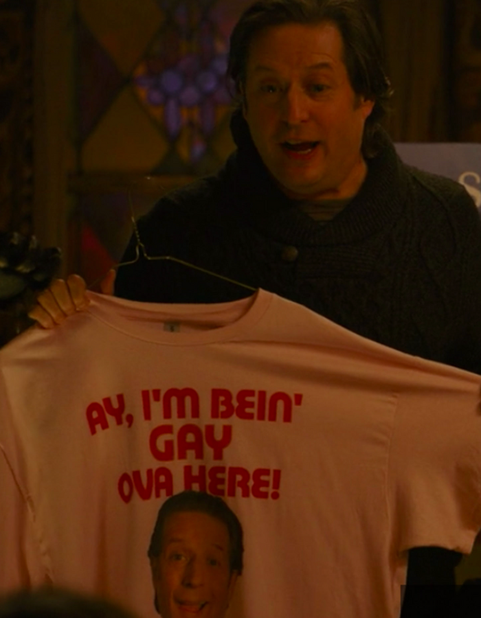 A man, the character Sean Rinaldi played by Anthony Atamanuik, is holding up a pink t-shirt with the text "AY, I'M BEIN' GAY OVA HERE!" and a picture of his face in the center from the comedy horror mockumentary fantasy TV series "What We Do In The Shadows" about vampires living in Staten Island.