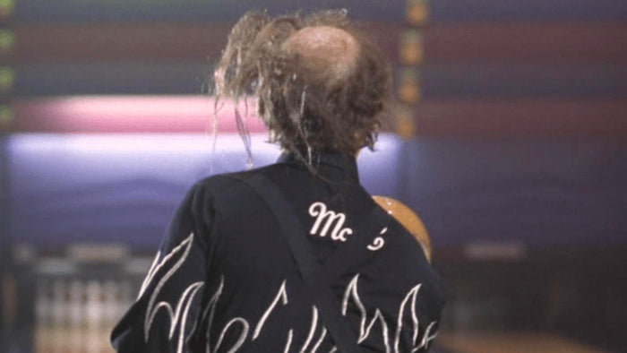 a person with a shaved head wearing a black shirt with white flames on the back, standing in front of a colorful background.