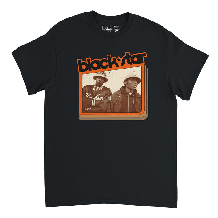 A vintage, souvenir "Black Star" t-shirt in the color black with the name of the iconic rap band outlined in the color orange on top of an image of the duo, Yasiin Bey and Talib Kweli, one wearing a Yankee jacket and the other a hoodie, both in hats. References the American hip hop due who named themselves after The Black Star Line shipping company, created by Marcus Garvey, and formed in Brooklyn, New York, with famous hits like "Respiration" and "Thieves in the Night".