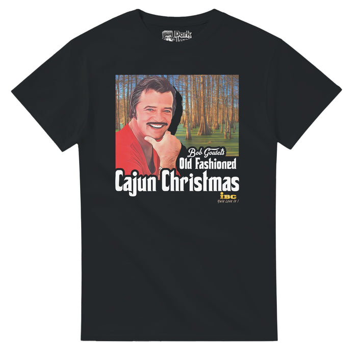 A vintage, black colored souvenir t-shirt that says "Bob Goulet's Old Fashioned Cajun Christmas" and "IBC, Yule Love It!" under an image of a man with a mustache and a red shirt in front of a forest. References the fictional holiday special featuring Robert Goulet on a raft, singing Christmas carols in a swamp with alligators chasing him from the 1988 dark satire movie Scrooged, a fresh take on Christmas Carol, which starred Bill Murray as TV executive Frank Cross as well as Karen Allen & John G