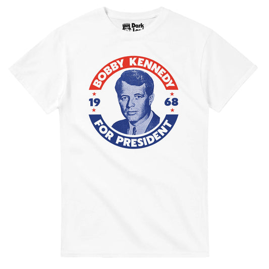 A white colored 80s style t-shirt with a red, white, and blue campaign sticker like graphic of Robert F. Kennedy's face in a blue tint, surrounded by the words "BOBBY KENNEDY FOR PRESIDENt, 1968" with little red stars, and references RFK's run for the Democratic presidential nomination before his assassination in June of 1968, similar to his brother, JFK's, assassination. RFK was known for his role as a lawyer, politician, the 64th United States attorney general, and US Senator from New York.