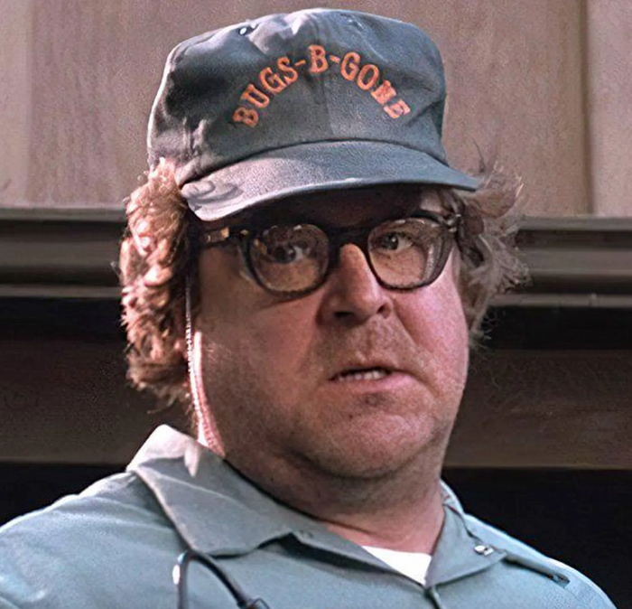 A closeup of character, Derbert McClintock, the exterminator played by actor John Goodman, wearing a sea green hat that says "BUGS-B-GONE" in orange text, glasses on his face, from the horror comedy thriller film Arachnophobia.