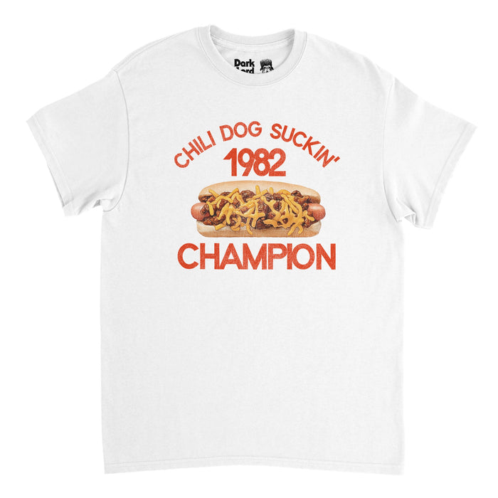 A retro, souvenir t-shirt in the color white that says "Chili Dog Suckin' 1982 Champion" with the image of a hotdog covered in chili and cheese inside a bun. References the iconic John Mellencamp (also known as John Cougar) hit chart topping song "Jack and Diane" in his album American Fool in 1982 in the genre of heartland rock country folk.