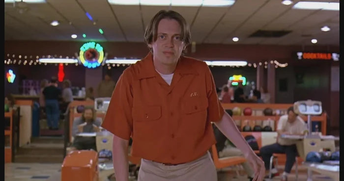 A man wearing an orange button up collared retro shirt in a bowling alley. From the 1998 screwball comedy movie "The Big Lebowski", worn by the character Donny, played by Steve Buscemi.