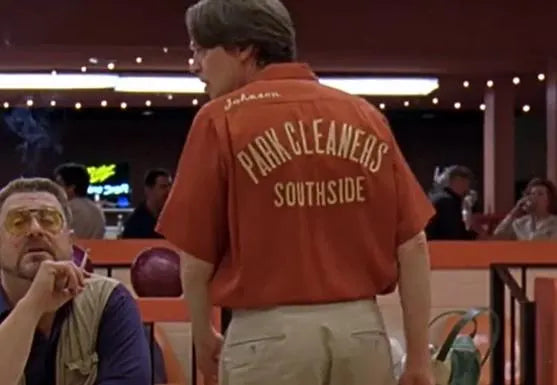 A man turned away, wearing an orange collared retro shirt that says "Park Cleaners Southside" and "Johnson" next to a guy smoking in a purple shirt in a bowling alley. References the comedy film "The Big Lebowski" from 1998 and was worn by Donny Kerabatsos (played by Steve Buscemi).