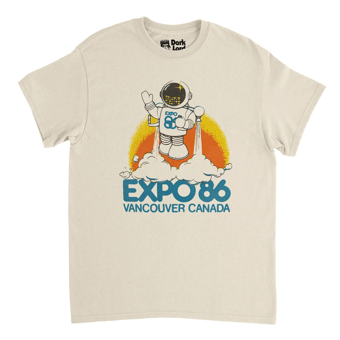 A retro, soft cream colored 80s style t-shirt that says "Expo 86 Vancouver Canada" in blue text, referencing the World Fair, called the "1986 World Exposition on Transportation and Communications". Includes an image of a robot in a spacesuit waving, taking off using jet propellers pushing white smoke out of them as he flies against the background of yellow and orange colors, referencing "Expo Ernie", the mascot of Expo '86.