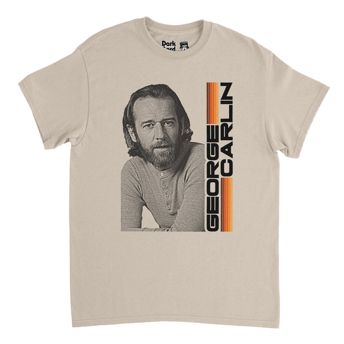 An off-white, sand colored vintage 80s style t-shirt that says GEORGE CARLIN in bold, capitalized black letters, next to lines of purple, orange, and yellow, and an image of George Carlin in black and white, the well known comedian, social critic, author, and actor who frequented The Tonight Show Starring Johnny Carson and was the host of the first ever Saturday Night Live show and created The George Carlin Show.
