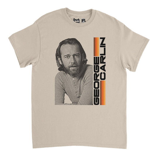 An off-white, sand colored vintage 80s style t-shirt that says GEORGE CARLIN in bold, capitalized black letters, next to lines of purple, orange, and yellow, and an image of George Carlin in black and white, the well known comedian, social critic, author, and actor who frequented The Tonight Show Starring Johnny Carson and was the host of the first ever Saturday Night Live show and created The George Carlin Show.