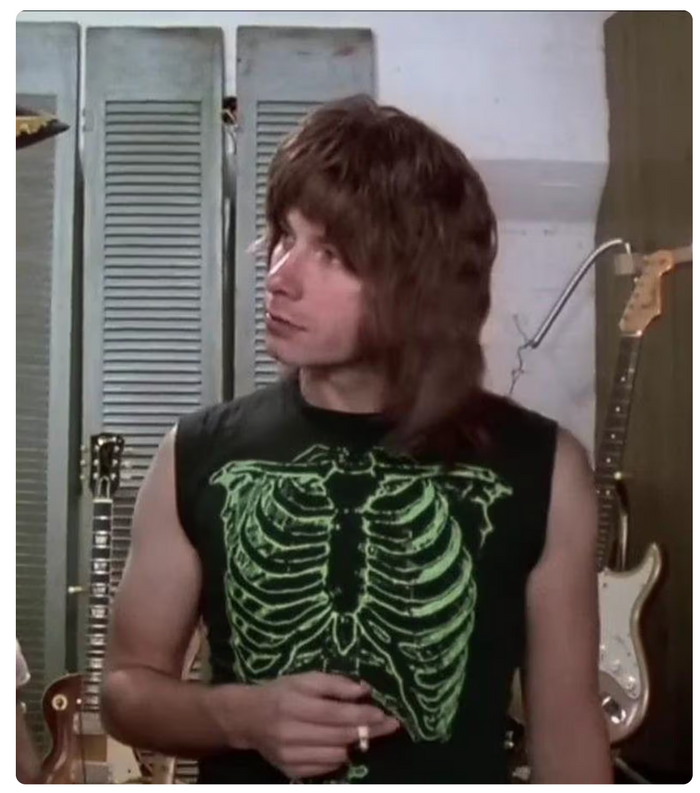 The character Nigel Tufnel, played by the actor Christopher Guest in the 1984 classic comedy mockumentary movie "This Is Spinal Tap". He has a long shag haircut and is wearing a black sleeveless shirt with a neon green skeleton graphic design, referred to as the Green Skeleton shirt, standing in front of a wall with guitar racks.