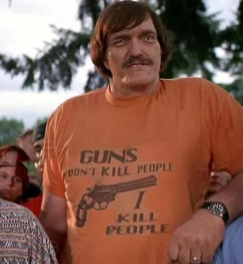 A man wearing a retro orange t-shirt with the image of a gun that says "Guns don't kill people I kill people". References the iconic 1996 movie "Happy Gilmore", worn by the character Mr. Larson played by Richard Dawson Kiel.