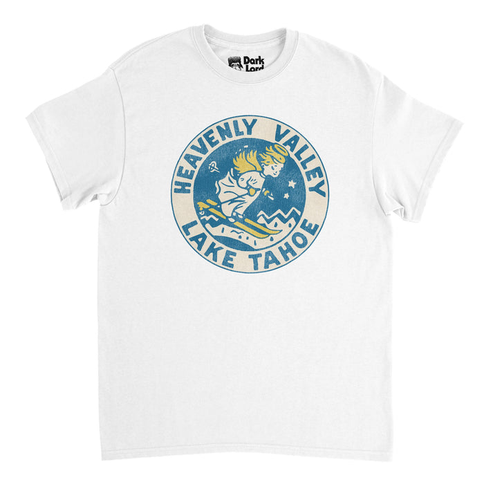 A vintage, souvenir Heavenly Mountain Resort t-shirt in the color white that says "Heavenly Valley Lake Tahoe" in blue letters inside a circle, surrounding a retro logo of the ski and snowboarding resort of a young girl skiing down hill with golden skis, golden angelic wings, golden hair, and a halo on top of her head, mountain in the background representing the Sierra Nevadas in both California and Nevada.