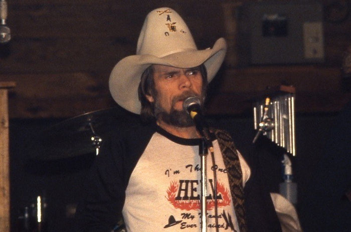 A man, iconic artist and singer Johnny Paycheck, wearing a cowboy hat and a t-shirt that says "I'm the only HELL my mama ever raised" with flames around the word 'hell' - referencing his famous 1977 hit song 'I'm the Only Hell (Mama Ever Raised)' - standing onstage and singing into a microphone