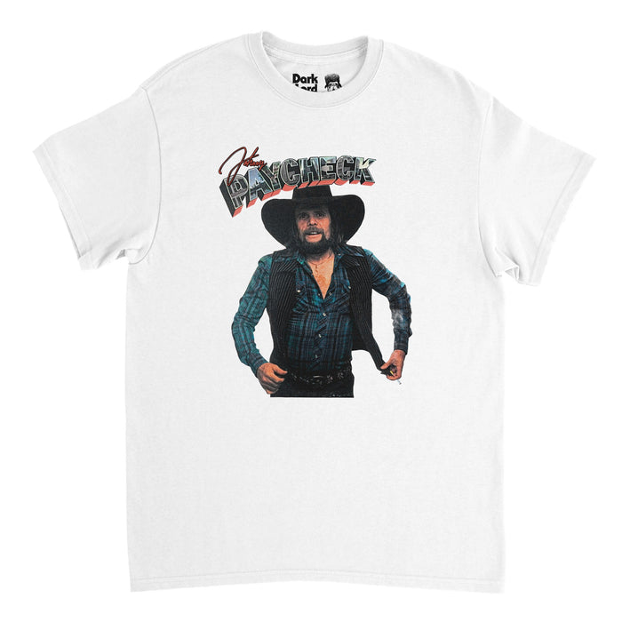 A vintage, 90s style t-shirt in the color white which spells "Johnny" in red cursive letters and "Paycheck" in bubble block letters that include city landscape images above the picture of Johnny Paycheck, a singer, songwriter, and founder of the outlaw country music movement, wearing a blue plaid flannel, striped vest, black cowboy hat, with a large beard. Was known for popular hit songs like "Take This Job and Shove It", "She's all I Got", "Someone to Give My Love To", and "Outlaw's Prayer".