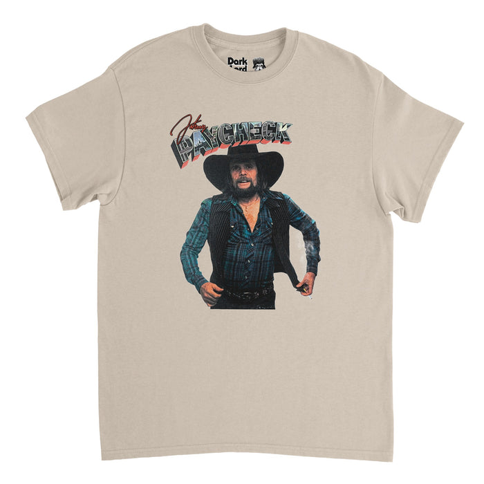 A retro, 70s style sand colored off-white t-t-shirt that says "Johnny" in red cursive and "Paycheck" in large block letters filled in with images of a city. Includes an image of the iconic country singer, songwriter, and founder of the outlaw country music movement named Johnny Paycheck, wearing a blue plaid flannel, striped vest, black cowboy hat, with a large beard. Had famous hit songs like "Take This Job and Shove It", "Old Violin", and "Outlaw's Prayer".