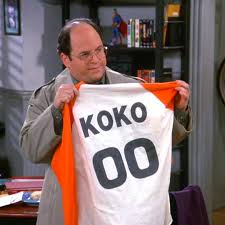 A man, the character George Costanza played by actor Jason Alexander, is holding up a red and white raglan style retro shirt with the name "KOKO" and number "00" on it. From the iconic American television sitcom "Seinfeld".