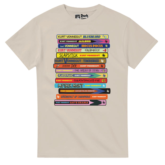 A sand colored retro t-shirt with the image of stack of famous Kurt Vonnegut books, their spines showing in various colors, including Bluebeard, Jailbird, Hocus Pocus, Slapstick, Timequake, Deadeye Dick, The Sirens of Titan, Player Piano, Slaughterhouse-Five, Mother Night, God Bless You, Mr. Rosewater, Breakfast of Champions, and Cat's Cradle. References the iconic, famous American author Kurt Vonnegut, Jr., known for his satire, dark humor, and science fiction novels, short stories, and plays.
