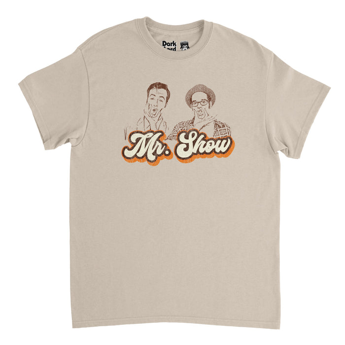 A retro, sand colored, vintage 80s style t-shirt that says "Mr. Show" in cursive letters outlined in brown and orange underneath the image of two men sketched, one wearing a checkered hat, plaid flannel, and glasses, the other a button up shirt, both holding their faces. References the iconic 1995 sketch comedy "Mr. Show with Bob and David" starring David Cross and Bob Odenkirk.
