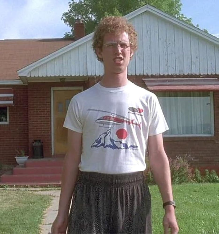 The character Napoleon Dynamite, played by actor Jon Heder, wearing a white t-shirt that says "Air Services International" and has a red helicopter. flying over the top of blue mountains and a red sun, as he stands in front of a brick house. From the retro feeling 2004 drama indie film "Napoleon Dynamite".