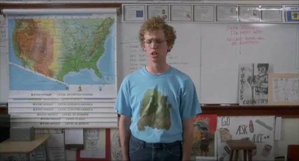 A young man with curly hair stands in front of a classroom with a map of the US, wearing a blue t-shirt with a graphic design of horses on a green/red background. From the comedic, iconic movie "Napoleon Dynamite", starring Jon Heder as the titular character.