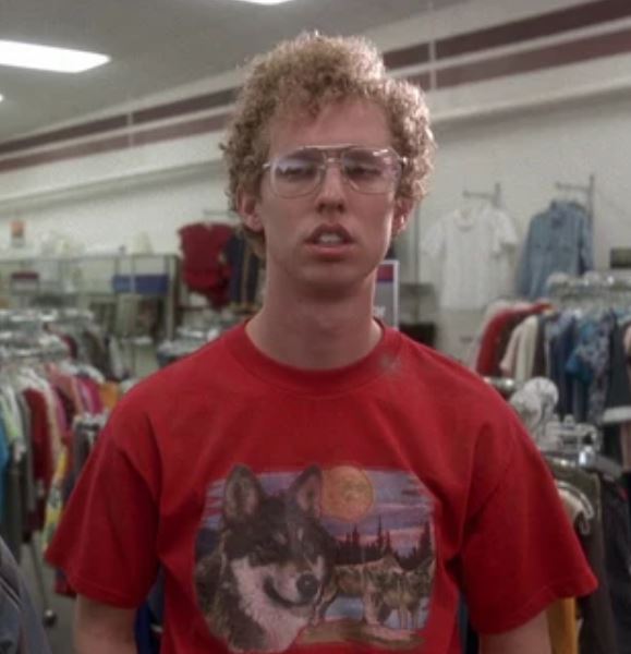 A man, character Napoleon Dynamite played by Jon Heder, is standing in a clothing department store, wearing a red t-shirt with the graphic of a wolf and a sunset, from the comedic teen indie film "Napoleon Dynamite".