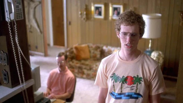 A man wearing a t-shirt with a graphic design of palm trees, a red sun, and blue water, standing in a room with a couch and a lamp, and another man at a desk. References the 2004 comedy indie movie Napoleon Dynamite starring Jon Heder as Napoleon Dynamite himself and Aaron Ruell as Kip Dynamite.