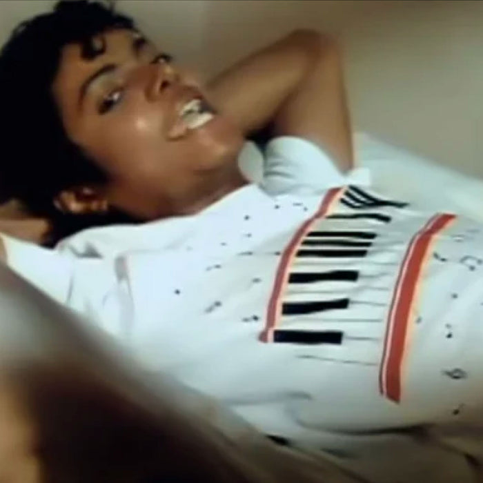 Michael Jackson lying down on a bed wearing a white short sleeved retro t-shirt with black music notes scattered throughout and piano keys bordered by red lines during his iconic music video for his 1982 hit song "Beat It".