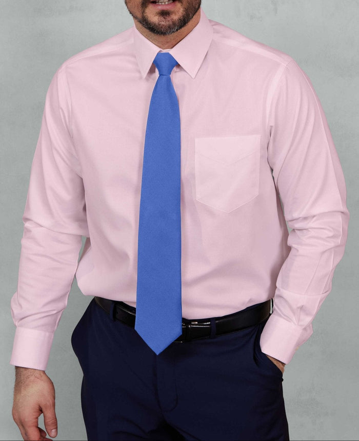 A man wearing a light pink long-sleeve dress shirt, a bright blue tie, and navy trousers, standing against a textured gray studio background.