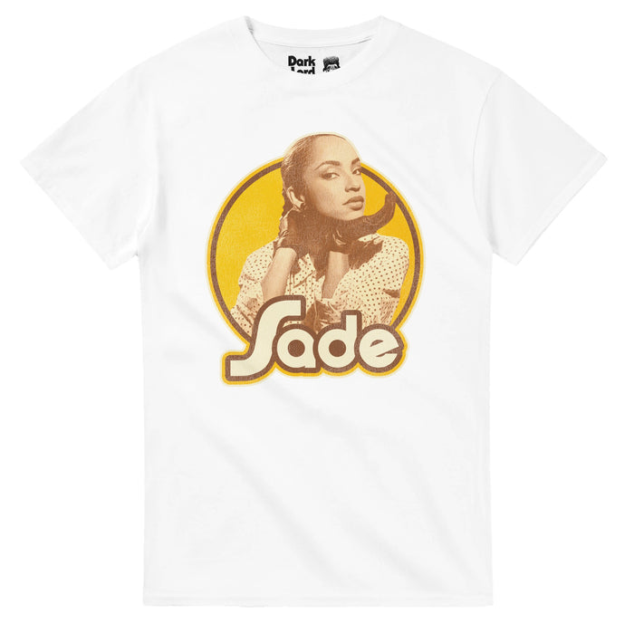 A vintage, 80s style white colored t-shirt that says "Sade" in light yellow bubble letters, outlined in brown, overlapping a circular yellow background with a sepia toned image of singer and songwriter Sade Adu popping out, wearing a polka dotted shirt, her hair pulled back, known for forming the iconic band Sade, also including Paul Denman, Stuart Matthewman, Andrew Hale, and Paul Cooke. Sade is known for hits like "Smooth Operator", "Your Love is King", "The Sweetest Taboo", and "Jezebel".