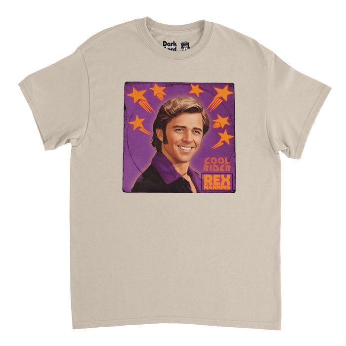 A retro, 80s style soft cream sand colored t-shirt with an image of actor Maxwell Caulfield playing character Rex Manning with his iconic sideburns, slicked back side part hair, and purple button up shirt on a background of purple with orange shooting stars. Says Cool Rider, referencing the cult classic film "Grease 2", and Rex Manning, referencing the coming of age movie "Empire Records".