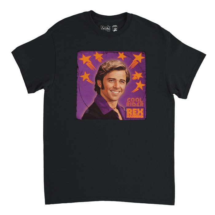 A vintage, souvenir black colored short sleeved t-shirt with a square purple background with orange shooting stars that says "Cool Rider Rex Manning" beside na image of a man - the character Rex Manning, played by Maxwell Caulfield - smiling with retro slicked back hair, sideburns, and a purple shirt. References the cult classic film Empire Records and the movie Grease 2.