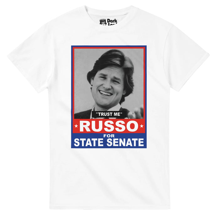 A retro, 80s style white colored short sleeved t-shirt that says the quote "Trust Me" and "RUSSO FOR STATE SENATE" with an image of actor Kurt Russell as Rudy Russo surrounded by rectangles of red and blue. References the iconic 1980 satirical comedy film "Used Cars" written and directed by Robert Zemeckis in which Russo enters politics and also stars Deborah Harmon, Gerrit Graham, Frank McRae, and Joseph P. Flaherty.