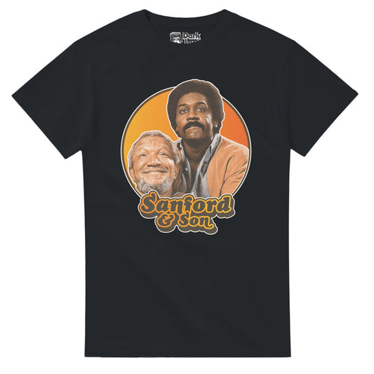 A 70s style souvenir black colored t-shirt that says "Sanford & Son" in orange and gray text underneath an image of iconic characters Fred G. Sanford and Lamont Sanford, played by actors Redd Foxx and Demond Wilson against an hombre yellow and orange background. References the 1972 hilarious sitcom television series "Sanford and Son" also starring Whitman Mayo, LaWanda Page and Don Bexley, based on the British sitcom "Steptoe and Son".
