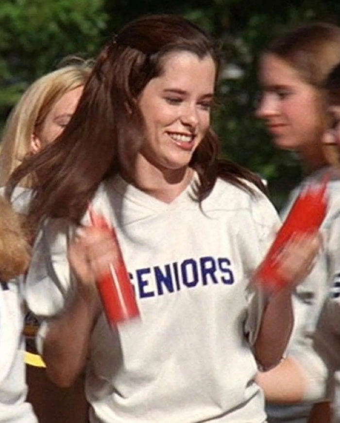 A young woman with long brown hair, high school senior Darla Marks, played by actress Parker Posey, is smiling and holding two ketchup bottles, wearing a retro white shirt with the word "SENIORS" written on it in navy blue, from the comedy indie film "Dazed and Confused".
