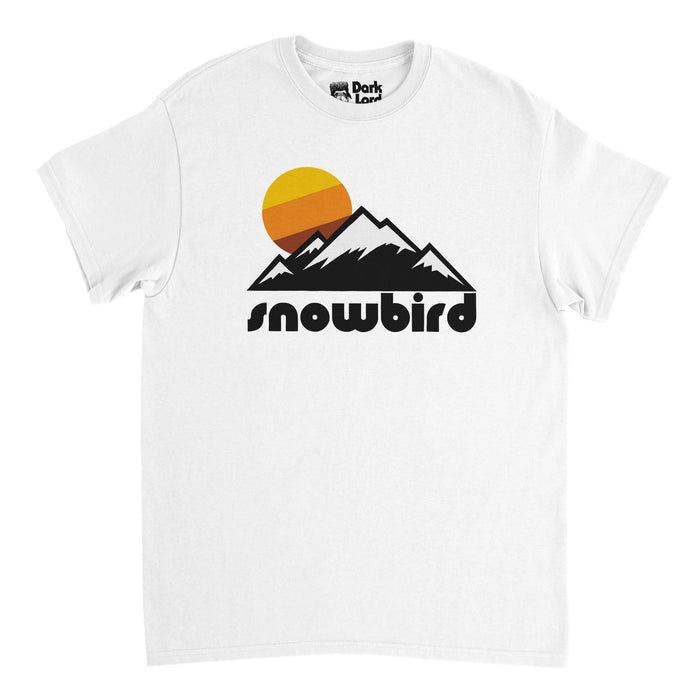 A vintage, 80s style souvenir white colored t-shirt that says "Snowbird" underneath the image of a snow covered mountain range, a yellow, orange, and brown sun setting above its peaks. References Snowbird Ski and Summer Resort near Salt Lake City, Utah in the Wasatch Range of the Rocky Mountains, with stunning skiing trails and hiking opportunities.
