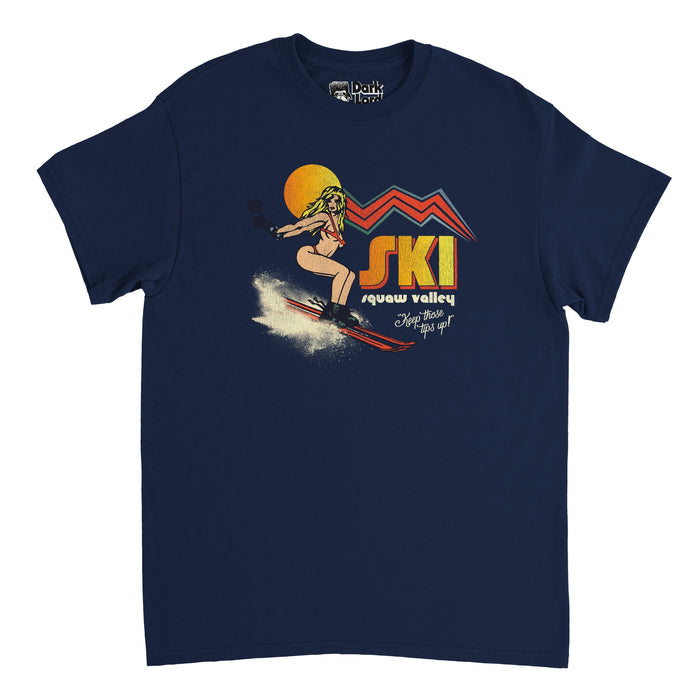 A retro, navy colored 80s style t-shirt that says "Ski Squaw Valley" with hombre orange to yellow letters, with the slogan "Keep those tips up!" Includes the image of a bikini clad girl skiing on red skis, snow flying up behind her, an orange/yellow sun behind her blonde head, and comic book like decorations resembling mountains, referencing Squaw Valley Ski Resort in California, now known as Palisades Tahoe, in the Sierra Nevadas.