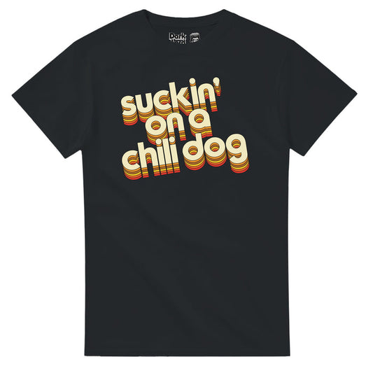 A retro, black colored 70s style souvenir short sleeved t-shirt that says "suckin' on a chili dog" in light yellow bubble letters with shadows of multiple shades of brown, yellow, and orange, highlighting the letters. References the lyrics "Suckin' on a chili dog outside the Tastee Freez, Diane's sittin' on Jacky's lap" from the iconic chart-topping hit song "Jack & Diane" by famous rock and folk singer John 'Cougar' Mellencamp.