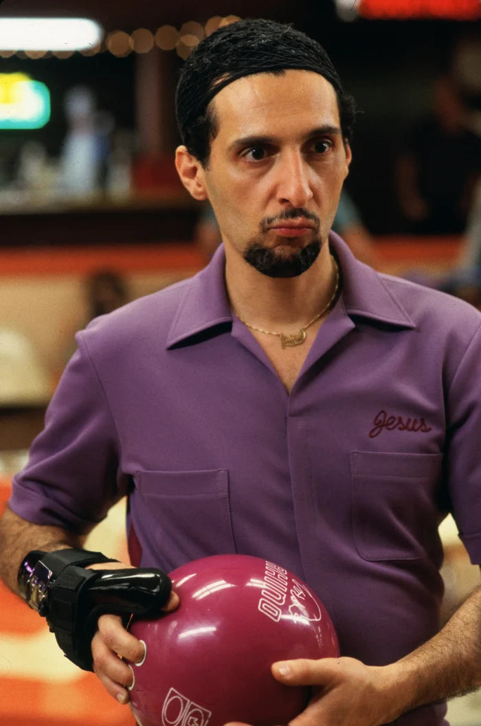 A man, Jesus Quintana played by John Turturro in the comedy film "The Big Lebowski", wearing a purple bowling jumpsuit inscribed with "Jesus" in red letters on the top left. He's holding a pink/red bowling ball, his hand covered by a wrist guard, and is determined, ready to bowl.