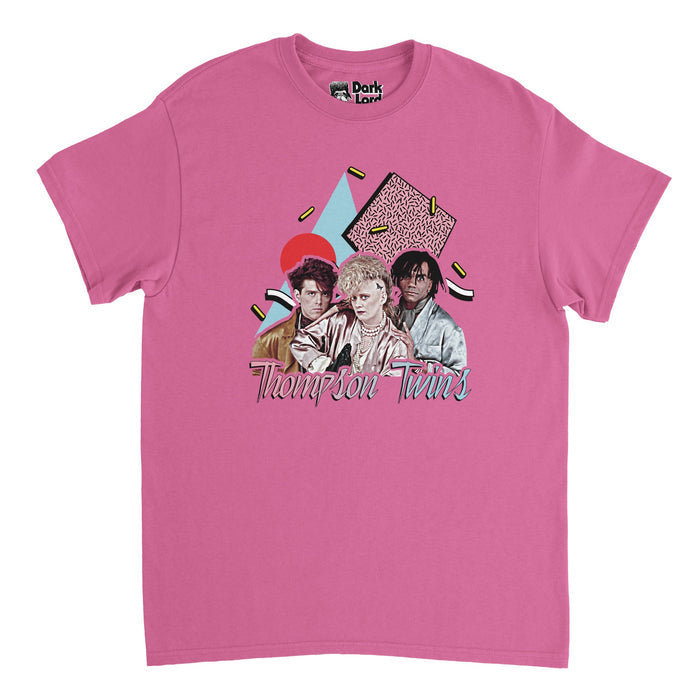 A bright pink 80s style souvenir t-shirt that says the name "Thompson Twins" in hombre pink and blue colors and includes an image of the three famous band members: Tom Bailey, Joe Leeway, and Alannah Curie in iconic, rocker outfits, over a background of shapes and blocks in red, blue, pink, and yellow colors. References the English pop band originally formed in Sheffield, originally a new wave music group, with famous album "Into the Gap" and songs like 'Make Believe" & "Lies".