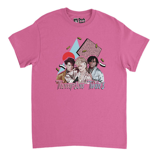 A bright pink 80s style souvenir t-shirt that says the name "Thompson Twins" in hombre pink and blue colors and includes an image of the three famous band members: Tom Bailey, Joe Leeway, and Alannah Curie in iconic, rocker outfits, over a background of shapes and blocks in red, blue, pink, and yellow colors. References the English pop band originally formed in Sheffield, originally a new wave music group, with famous album "Into the Gap" and songs like 'Make Believe" & "Lies".