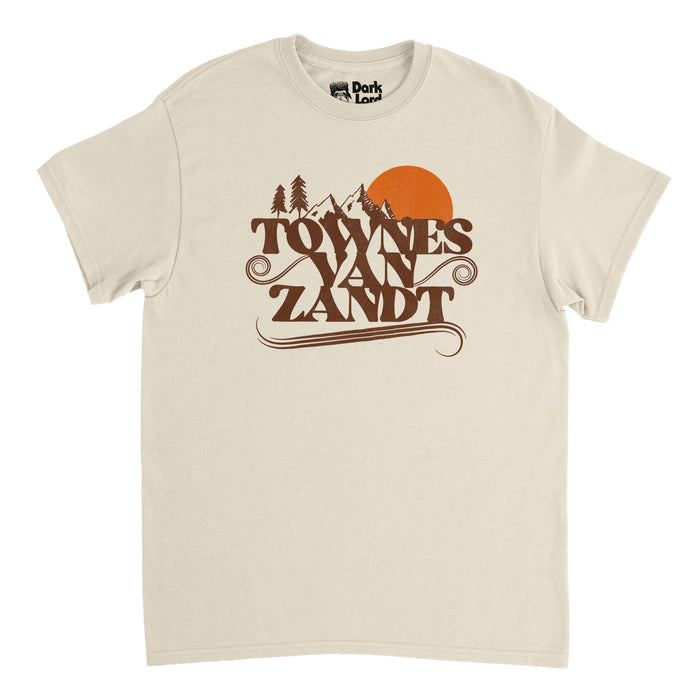 A soft cream colored, vintage white short sleeved 80s style t-shirt that says "Towns Van Zandt" in brown letters, swirls emanating from the sides and bottoms, and a couple tall mountain peaks next to a setting bright orange sun and pine trees.