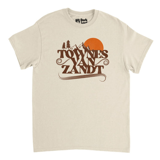 A soft cream colored, vintage white short sleeved 80s style t-shirt that says "Towns Van Zandt" in brown letters, swirls emanating from the sides and bottoms, and a couple tall mountain peaks next to a setting bright orange sun and pine trees.