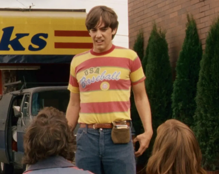 The retro image of a man, actor Jorma Taccone playing Kevin Powell, stands in a striped orange and yellow vintage t-shirrt that says "USA Baseball" in the iconic 2007 comedy drama film "Hot Rod", also starring Andy Samberg as Rod Kimble and Isla Fisher as Denise Harris.