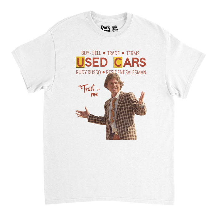 A vintage, 70s style white colored short sleeved t-shirt that says "Buy, Sell, Trade, Terms" above the title of iconic 1980 satirical film "Used Cars" and "Rudy Russo, Resident Salesman" above an image of Russo himself - portrayed by famous actor Kurt Russell - wearing a checkered brown suit and tie next to his popular quote, 'Trust me'.